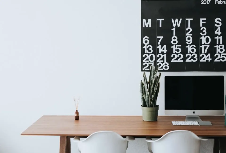 A wooden desk with a plant and a monitor against a white wall, a large wall calendar above.