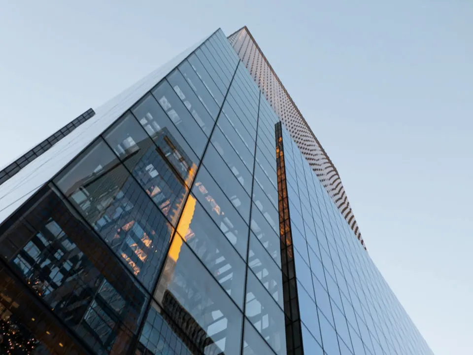 Looking up the sloping glass wall of a modern office tower against a pale sky.