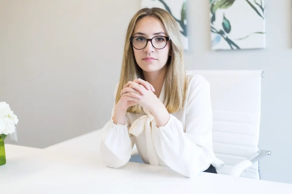 Illustrative portrait of a woman in a white blouse and glasses, seated at a white desk with her hands clasped.