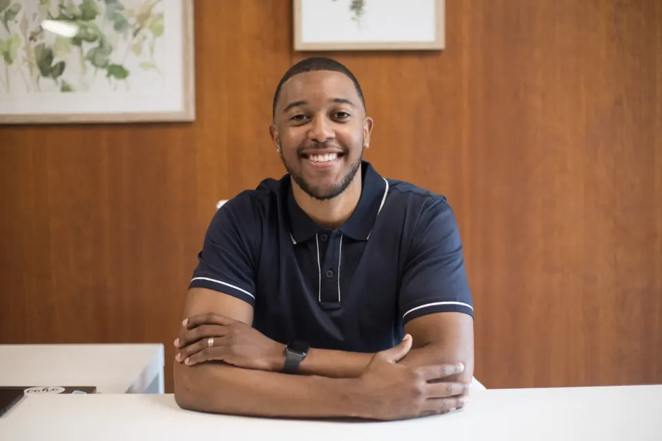 Illustrative portrait of a smiling man in a navy polo shirt, seated at a white desk in a wood-paneled room.