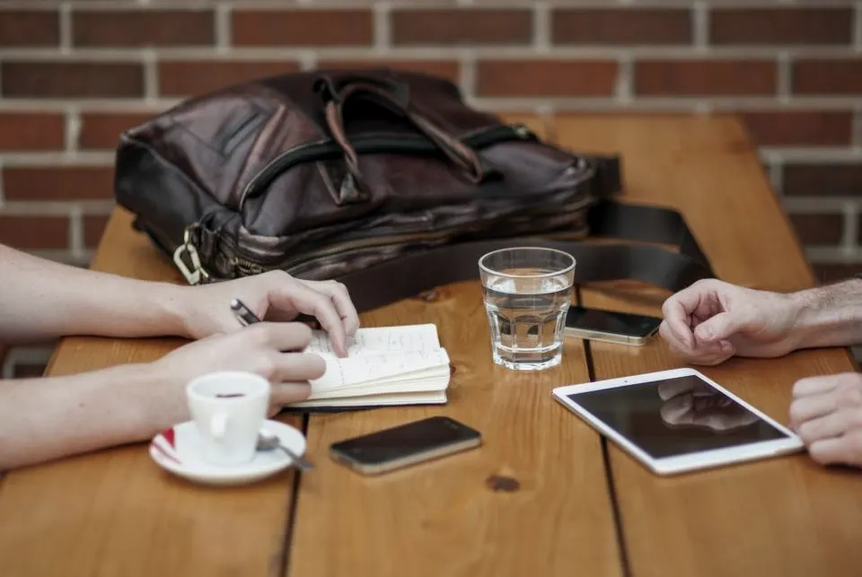 Hands holding pens over notebooks on a wooden table with coffee, a tablet and a leather bag.